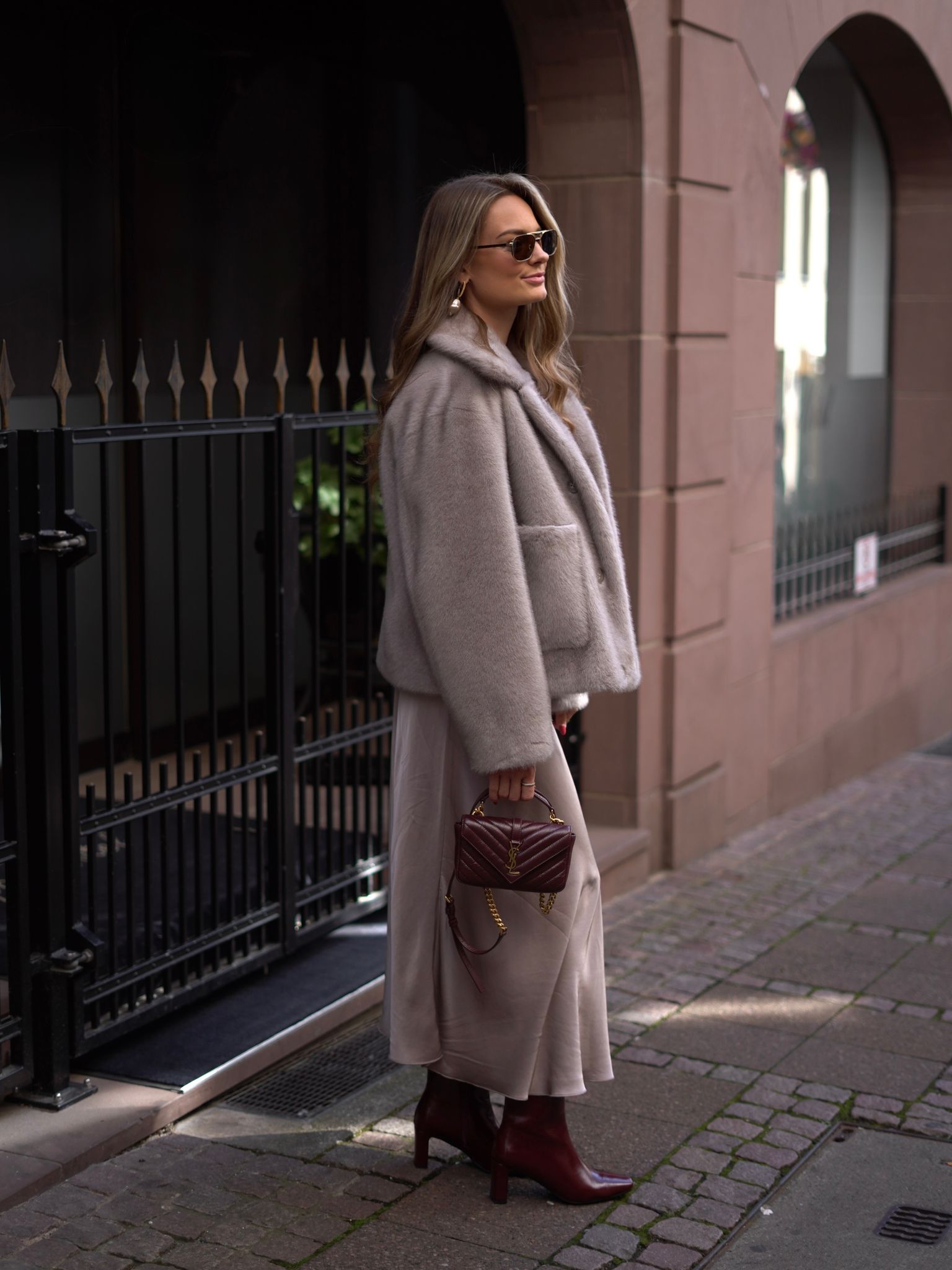 Frau in beige Mantel und Stiefeln vor rotem Backsteingebäude mit Handtasche und Sonnenbrille.