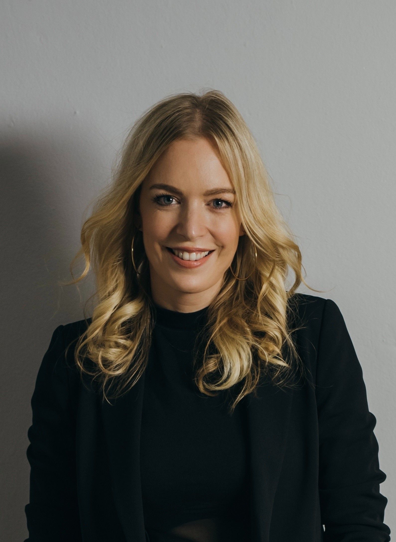 Lächelnde Frau mit blonden Locken in schwarzem Blazer vor hellem Hintergrund.