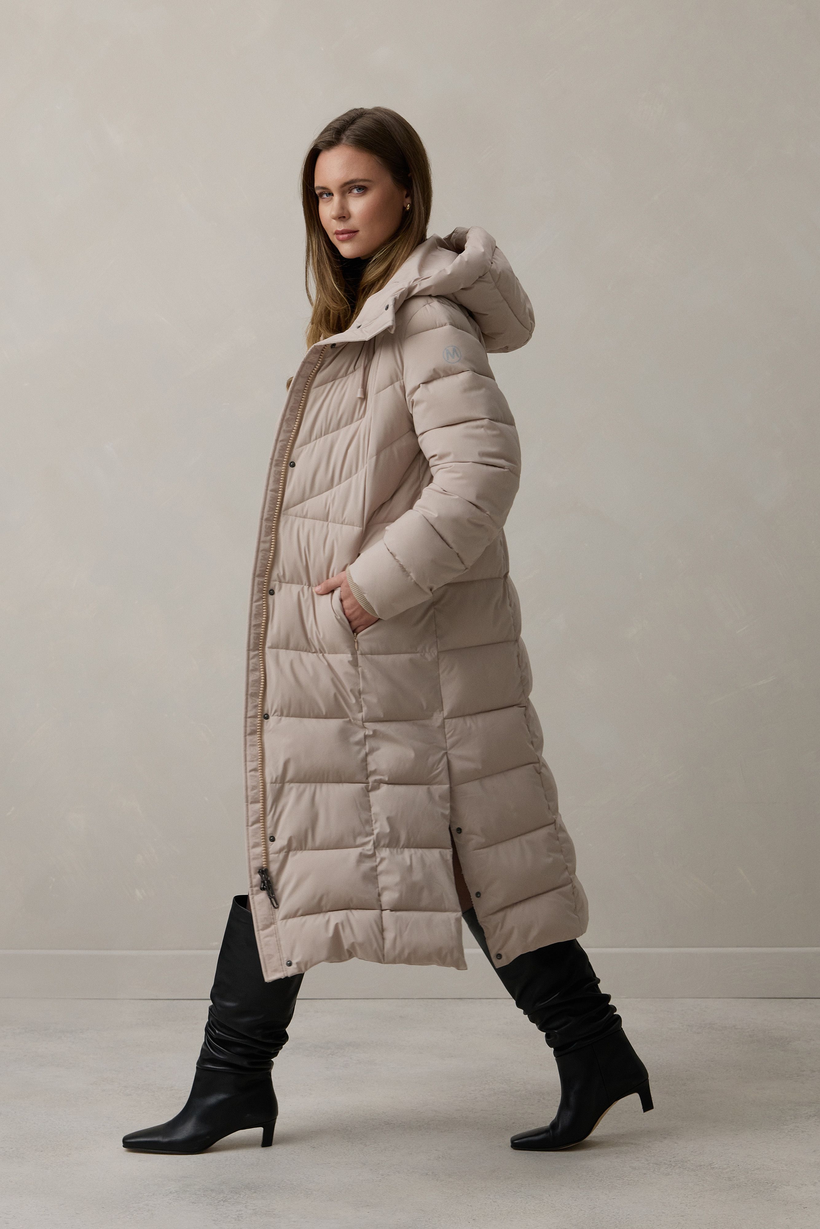 Frau trägt beige, langes Daunenmantel-Outfit mit schwarzen Stiefeln und lässiger Haltung.