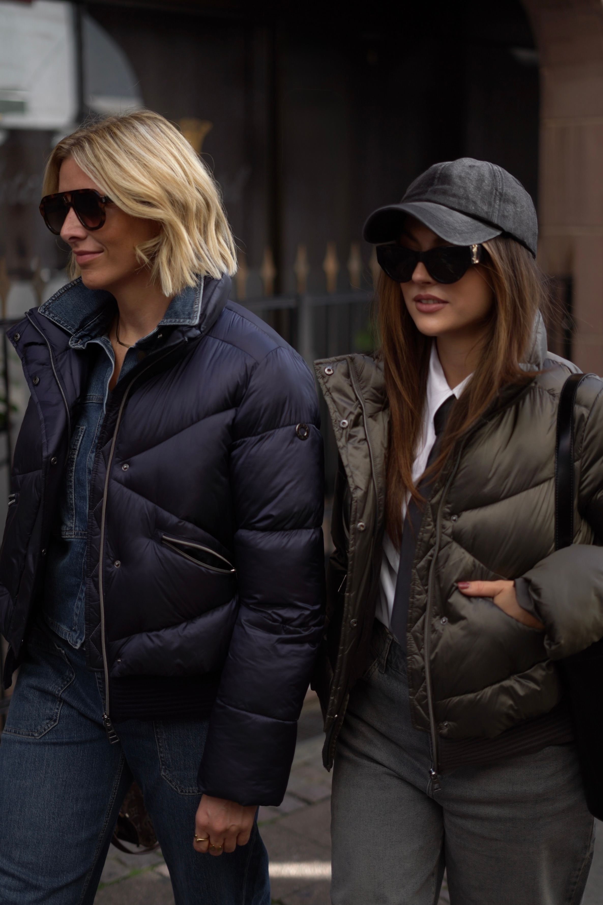 Zwei Frauen in stylischen Pufferjacken und Sonnenbrillen laufen nebeneinander auf der Straße.