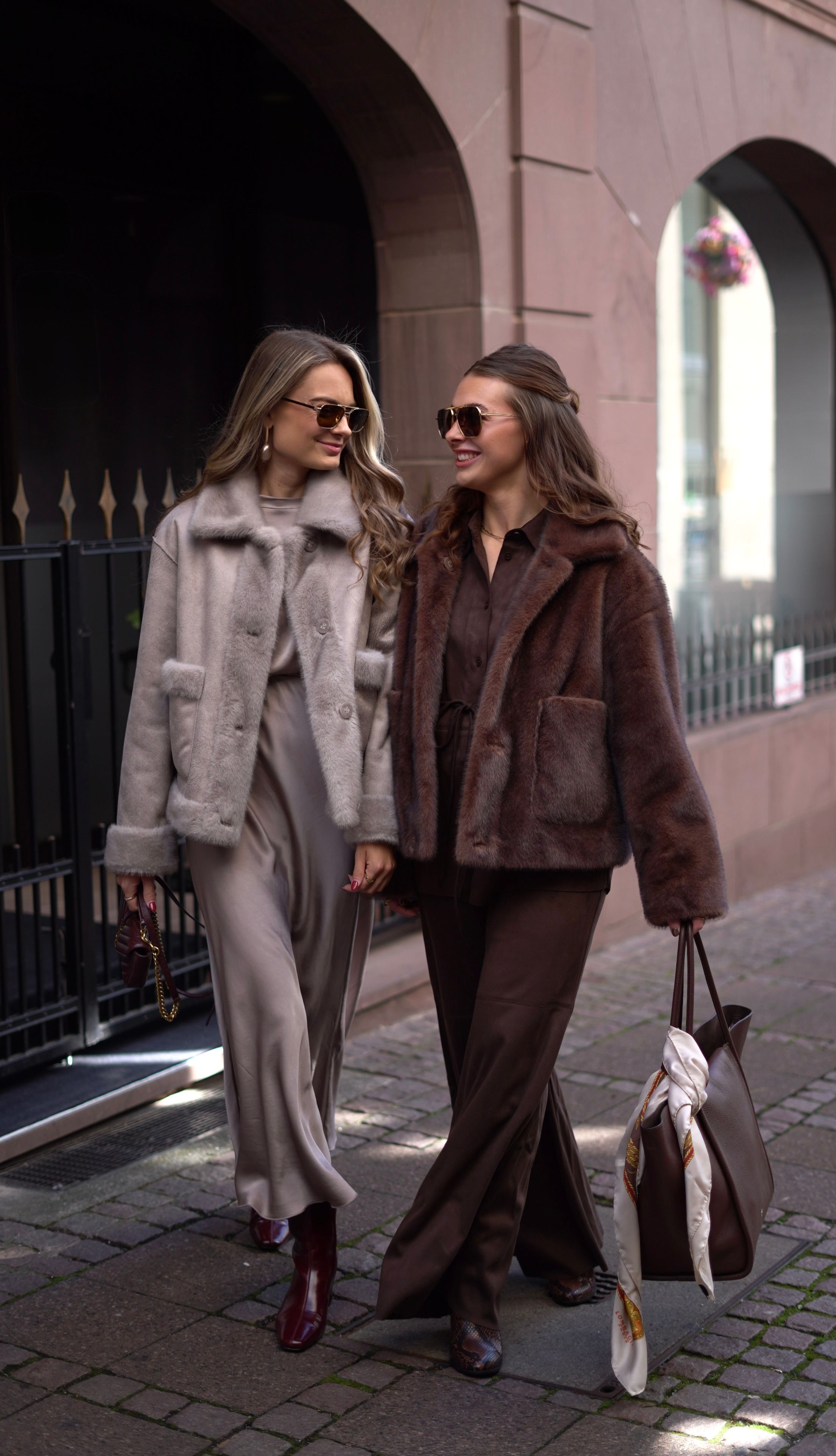 Zwei Frauen in eleganten Mänteln und Anzügen spazieren nebeneinander auf einer Straße.